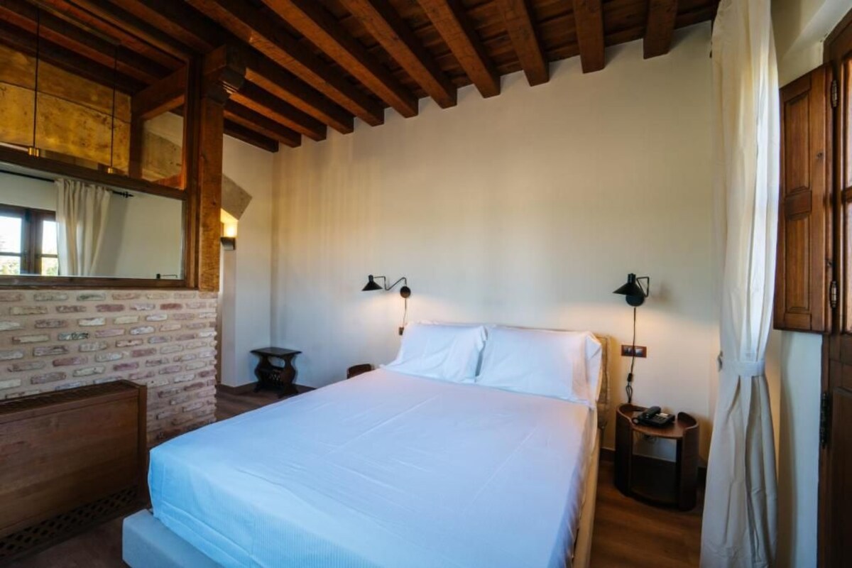 A bedroom with a large bed, wooden beams, brick wall, two bedside tables with lamps, and a mirror.