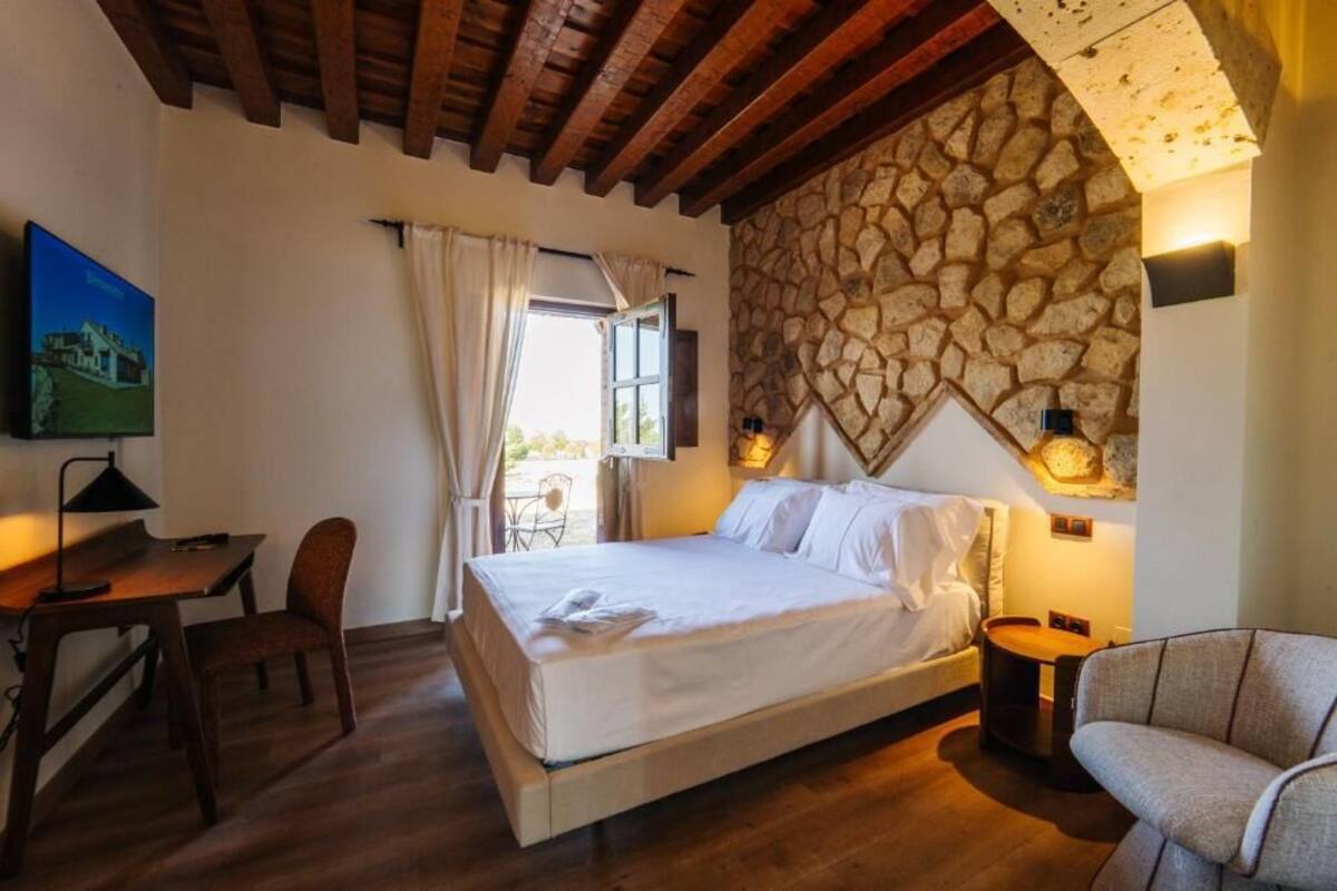 Une chambre à coucher avec un lit, un bureau, une chaise, un mur en pierre et une fenêtre avec des rideaux.
