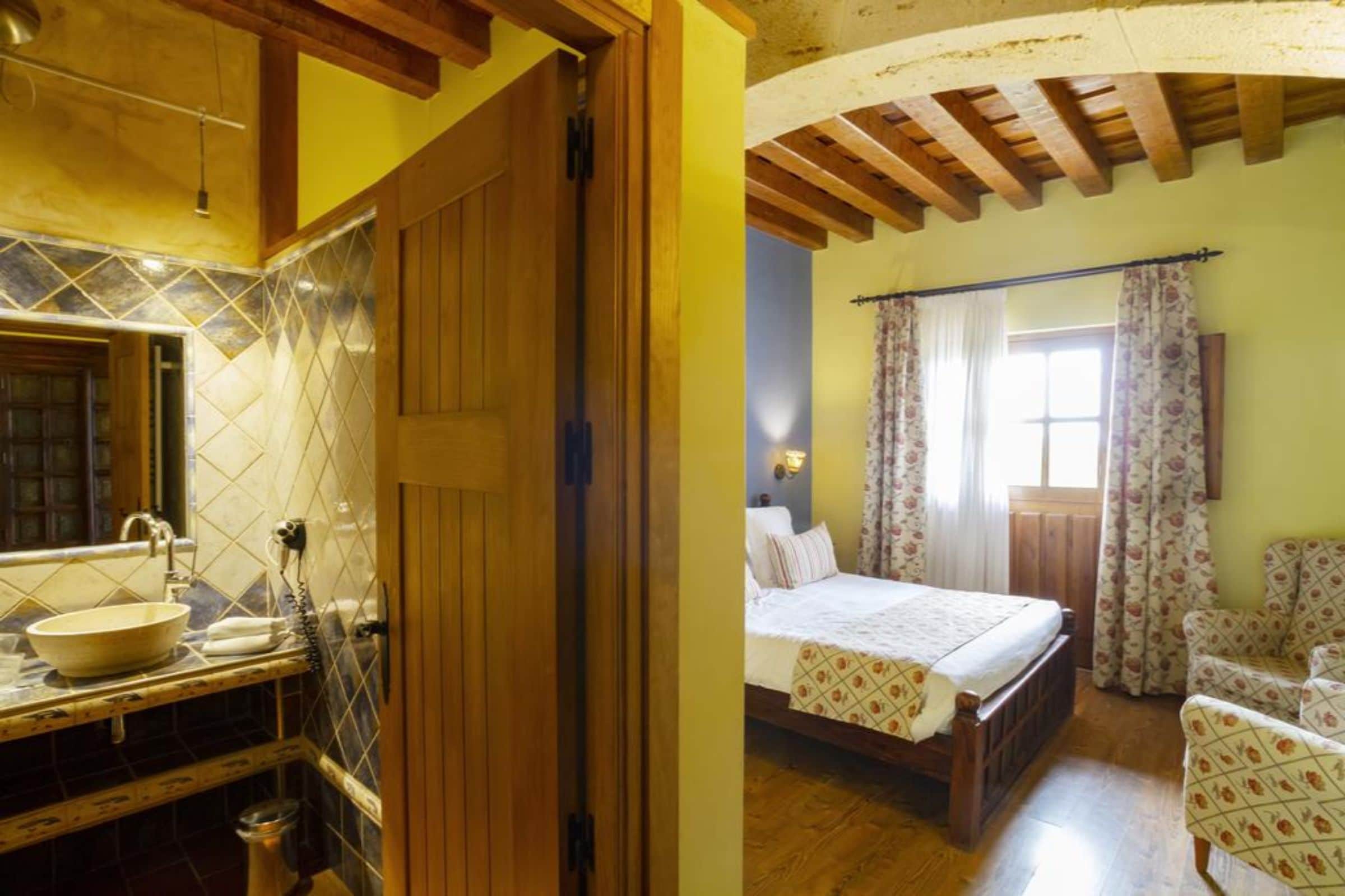 A room with a wooden ceiling, a bed with floral bedding, a chair, and a bathroom with a sink and mirror.