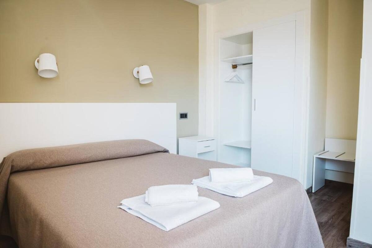 A hotel room with a bed, two wall-mounted lamps, a wardrobe, and a desk.