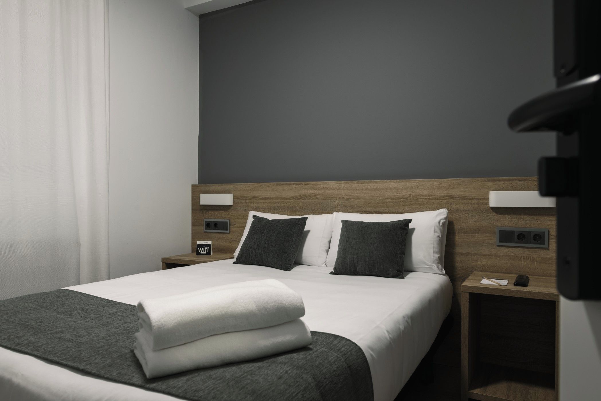 A modern hotel room with a neatly made bed, a wooden headboard, and a nightstand with a phone and a small lamp.