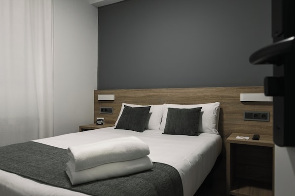 A modern hotel room with a neatly made bed, a wooden headboard, and a nightstand with a phone and a small lamp. - Alda Narón (Naron)