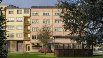 Un edificio residencial de varios pisos con balcones, un pequeño árbol y un coche rojo estacionado al frente.