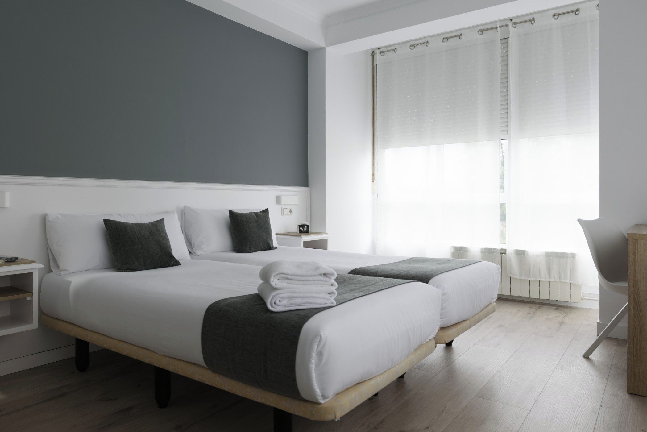 A modern hotel room with two beds, a nightstand, and a view of the outdoors through sheer curtains.