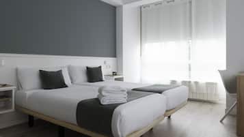 Quarto de hotel moderno com duas camas, uma mesinha de cabeceira e vista para o exterior através de cortinas translúcidas.