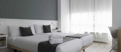 Habitación de hotel moderna con dos camas, una mesita de noche y vistas al exterior a través de cortinas translúcidas.