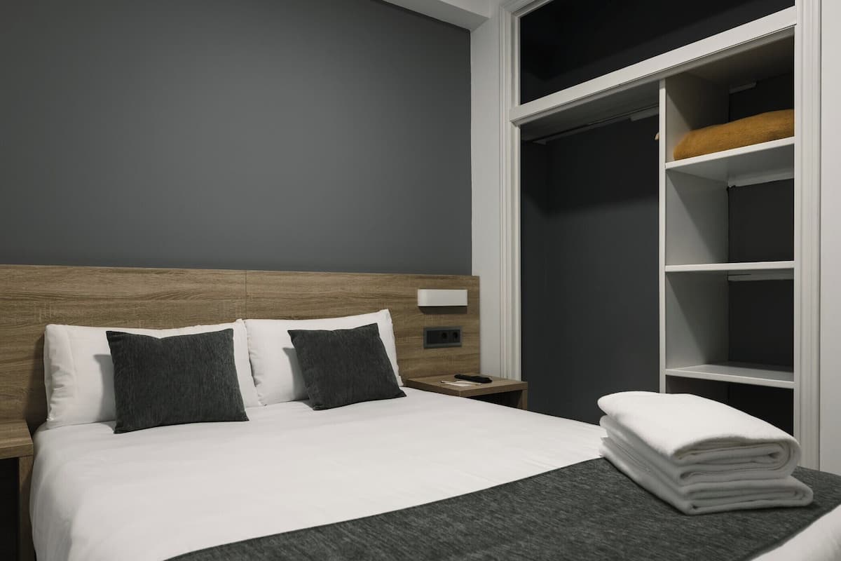 A neatly made bed with white linens and two dark pillows, a wooden headboard, a bedside table with a lamp, and a white closet with shelves.