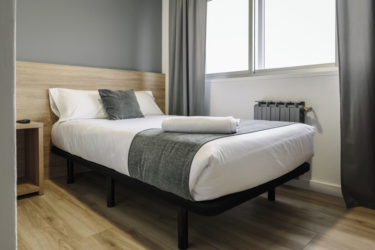 A modern bedroom with a wooden headboard, a neatly made bed with white linens and a grey throw, a bedside table, and a radiator.