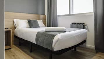 Quarto moderno com cabeceira de madeira, cama bem arrumada com lençóis brancos e manta cinza, mesa de cabeceira e radiador.