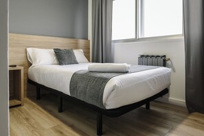 Un dormitorio moderno con cabecera de madera, cama con ropa de cama blanca y una manta gris, mesita de noche y radiador.