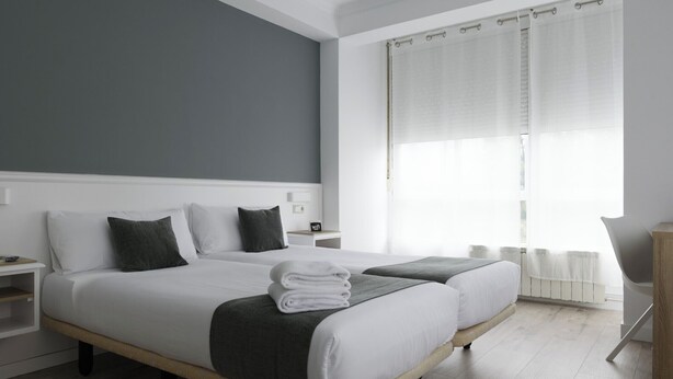 Ein modernes Hotelzimmer mit zwei Betten, einem Nachttisch und Blick ins Freie durch transparente Vorhänge.