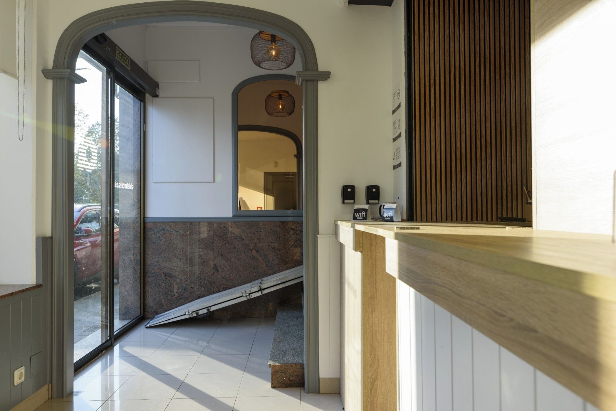 Un área de recepción con mostrador de madera, puerta de vidrio de entrada y una pequeña escalera que sube a un nivel superior.