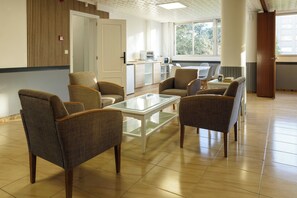 Habitación amplia con diseño moderno, que cuenta con una zona de estar con sillas y una mesa de vidrio, una kitchenette al fondo y ventanas grandes que ofrecen luz natural.