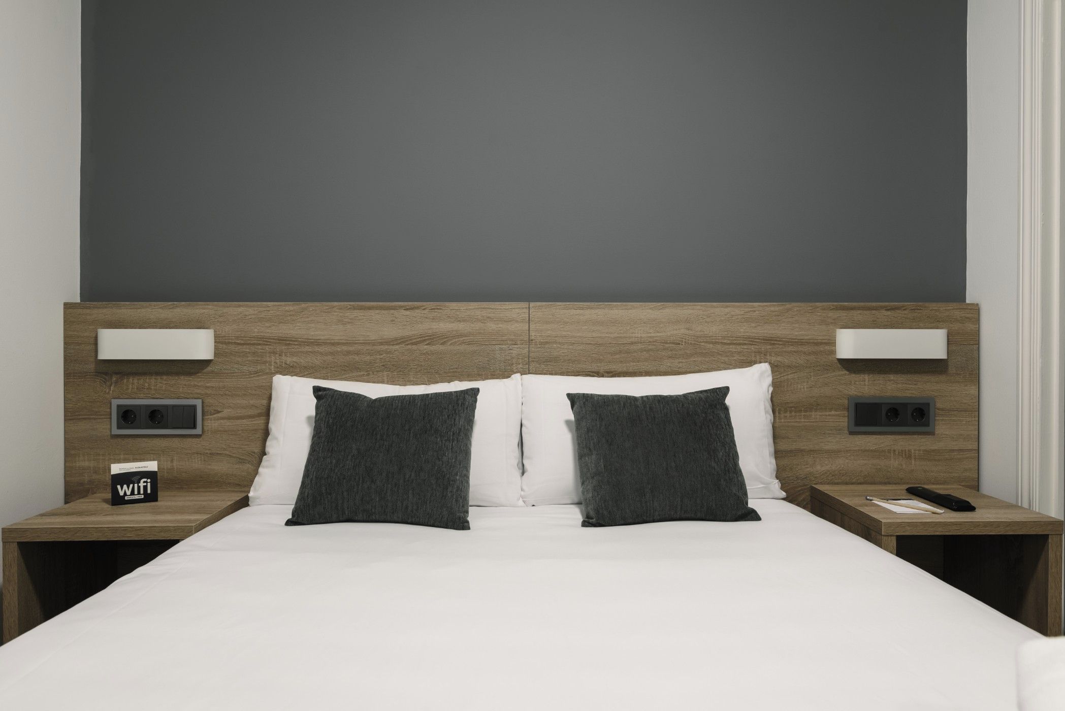 A hotel room with a wooden headboard, white bedding, two dark pillows, bedside tables with lamps, and a 'wifi' sign.