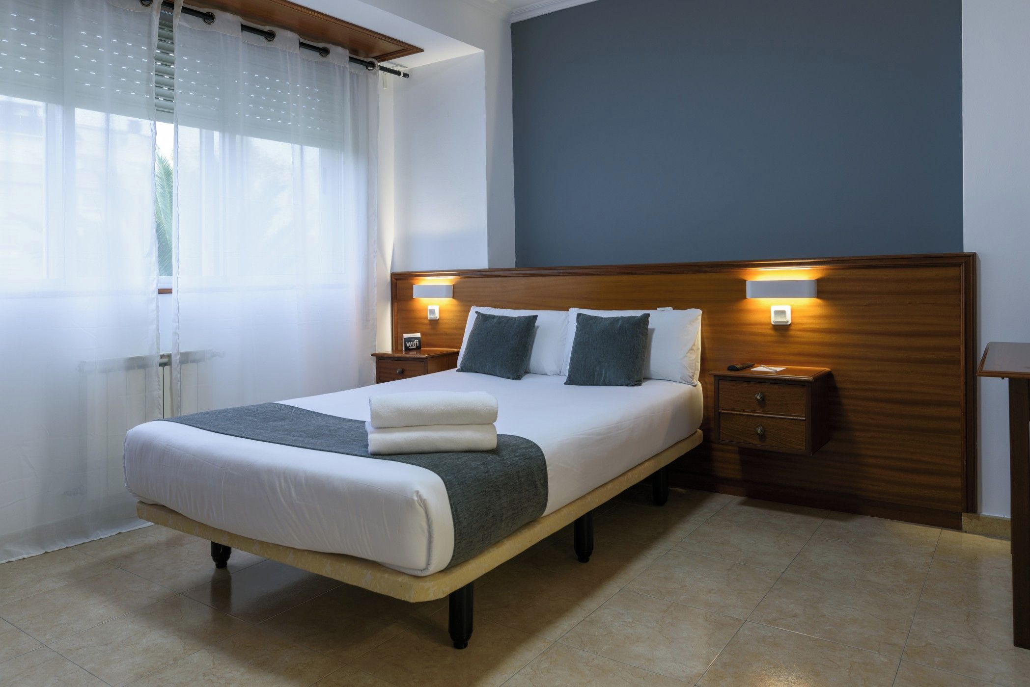 A neatly made bed with a wooden headboard, two bedside tables with lamps, and a window with sheer curtains.