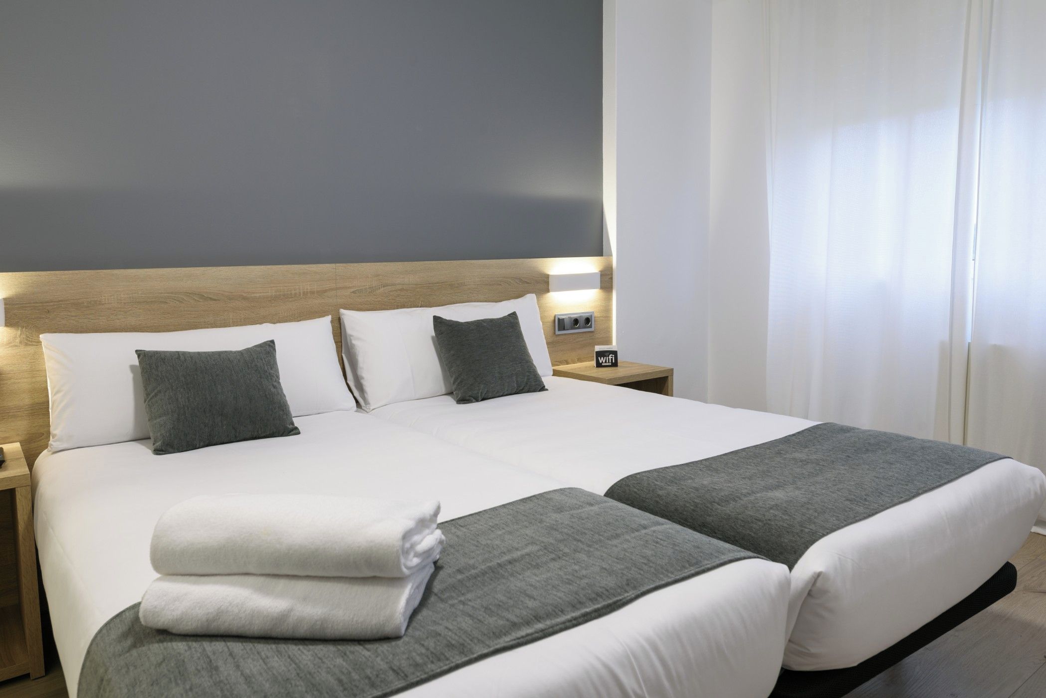 Cama doble con ropa de cama blanca y gris, cabecera de madera y mesita de noche con símbolo de Wi-Fi.