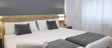 Cama doble con ropa de cama blanca y gris, cabecera de madera y mesita de noche con símbolo de Wi-Fi.