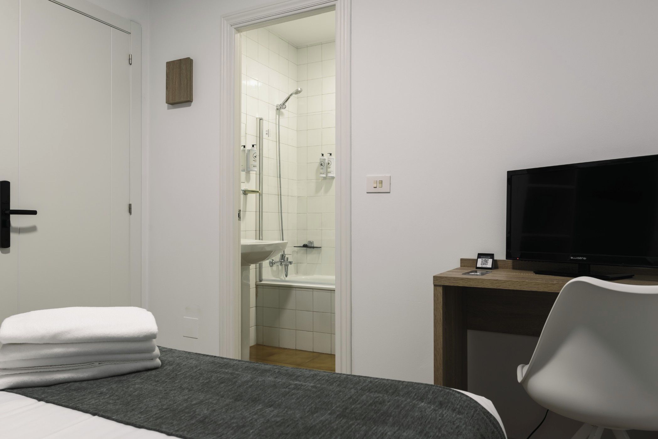 Habitación de hotel con cama, escritorio con televisor, baño con ducha y lavamanos, y una pila de toallas.