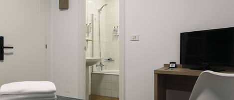 Habitación de hotel con cama, escritorio con televisor, baño con ducha y lavamanos, y una pila de toallas.