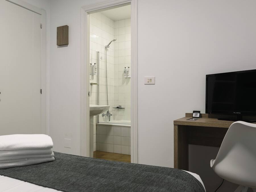 A hotel room with a bed, a desk with a television, a bathroom with a shower and sink, and a stack of towels.