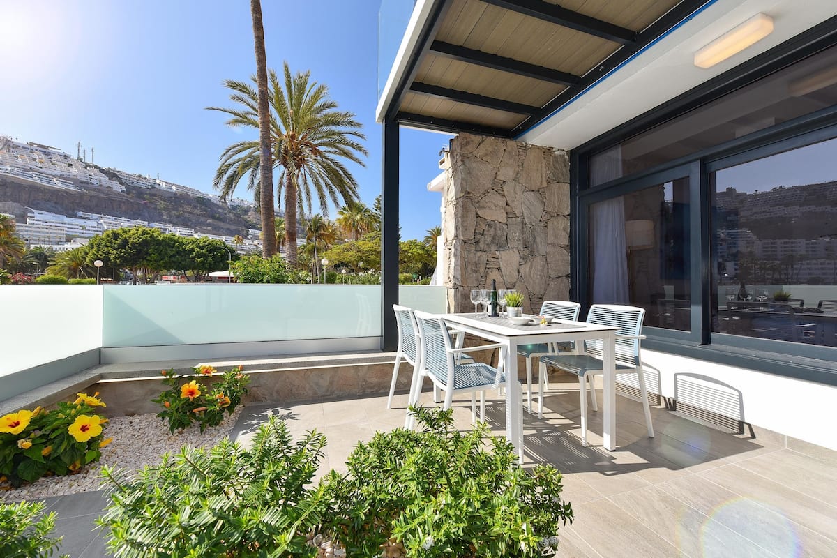 A modern outdoor patio with a stone wall, white furniture, and a view of a hillside.