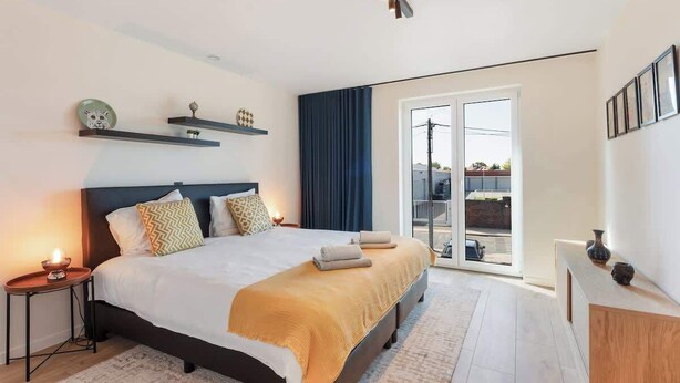 A modern bedroom with a large bed, a bedside table with a lamp, a window with a view, and a sliding glass door to the balcony. - Modern & Spacious with Wellness (Kortrijk)