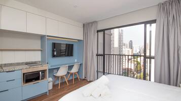 A modern hotel room with a bed, a small kitchenette, a seating area with chairs, and a large window with a city view.
