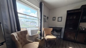 Ein Wohnzimmer mit zwei Sesseln, einem kleinen Tisch mit Pflanze, einem Fenster mit Vorhängen sowie einer Küchenzeile mit Schränken.