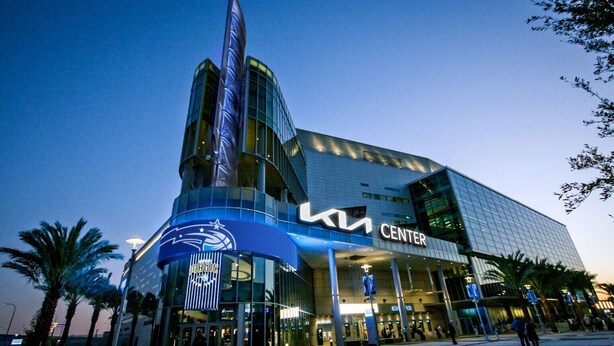 Ein modernes Gebäude mit einem auffälligen 'KIA CENTER' Schild, das eine geschwungene Glasfassade und eine hohe, zylindrische Struktur aufweist.