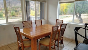 Un comedor con mesa de madera y seis sillas, ventanas amplias con persianas, y vista a un paisaje montañoso.