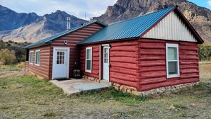 Una cabaña de troncos rojos con techo de metal, puerta blanca y chimenea. Está rodeada de un área cubierta de césped con montañas al fondo.