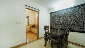 A dining area with a glass-top table and wooden chairs, a patterned tapestry on the wall, and a doorway leading to a kitchen. - Amazing Hridayam 2-bedroom bungalow in Panchgani (Panchgani)