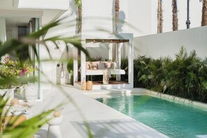 A modern poolside area with a lounge chair and a table, surrounded by lush greenery and a clear pool. - Mazari Villas (Bali)