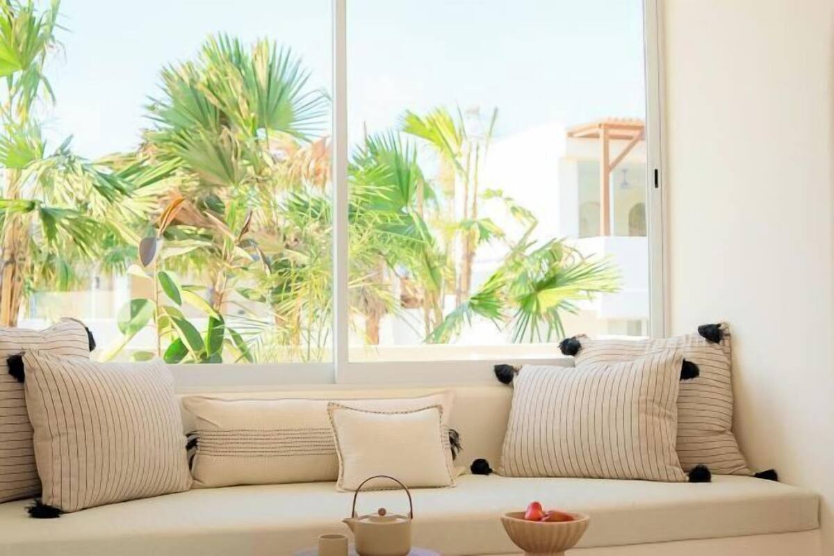 A sofa with striped cushions, a small table with a teapot and a bowl, and a view of palm trees through a window.