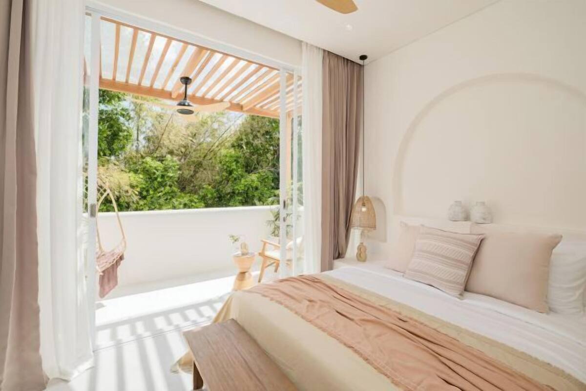 A bedroom with a bed, a bench, a lamp, a window with curtains, and a view of greenery outside.