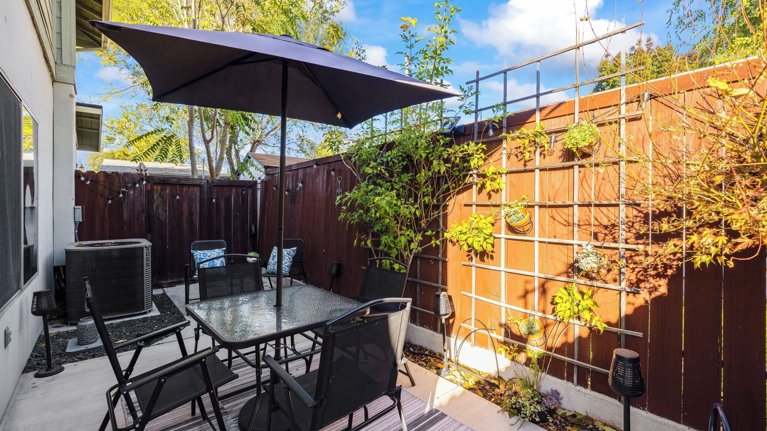 A patio with a table and chairs, a large umbrella, a wooden fence with climbing plants, and a metal trellis with potted plants.