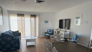 A modern living room with a sofa, a coffee table, a dining table with chairs, a TV, and a window with blinds. - Tranquil villa with private beach, 2br/2ba 3 pools, complex on beach!! Patio (Sarasota)