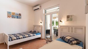 Quarto com duas camas de solteiro, uma mesinha e uma cadeira. Há um ar condicionado na parede e uma porta que dá para uma varanda.