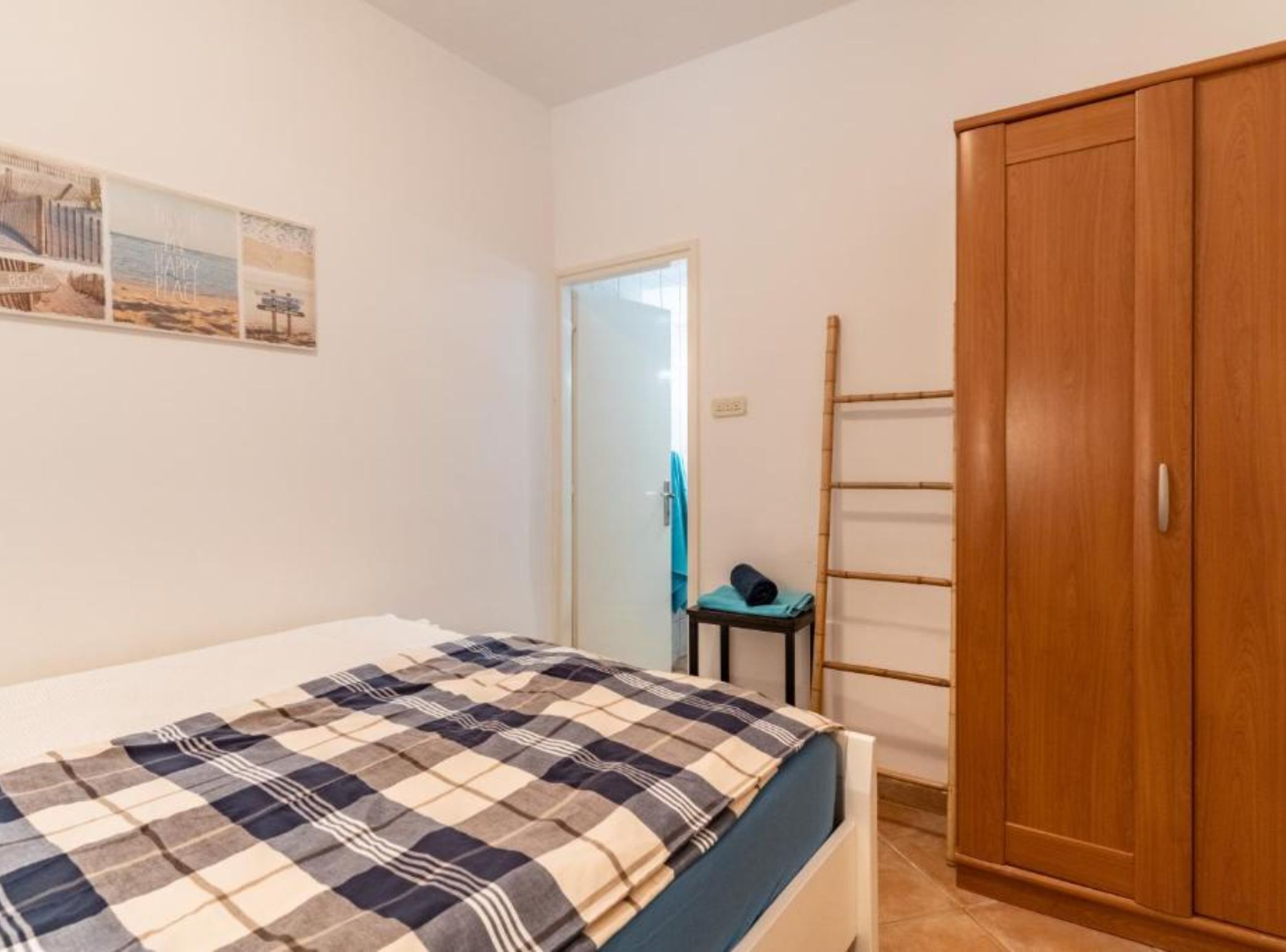 A bedroom with a bed, a wooden wardrobe, a small table with a towel, and a wall with a beach-themed poster.
