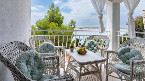 Un balcon agrémenté de meubles en osier, une table avec un panier de fruits et une vue sur la mer et la verdure.