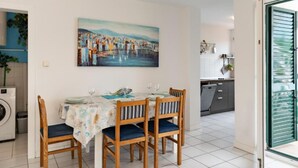 Un coin repas avec une table dressée pour quatre personnes, un tableau représentant un paysage côtier et une vue sur la cuisine et la buanderie.