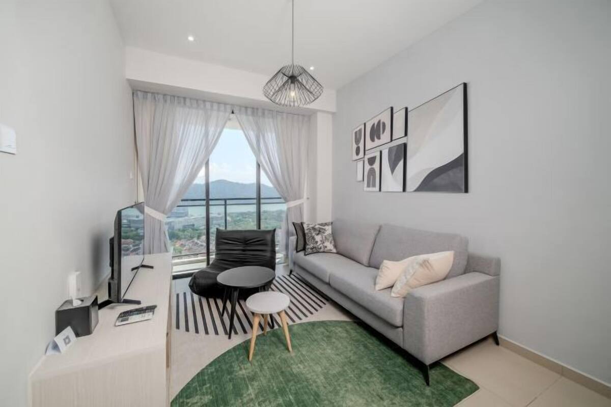 Een moderne woonkamer met een bank, een bureau met een computer, een klein rond tafeltje en uitzicht op het groen door grote ramen.