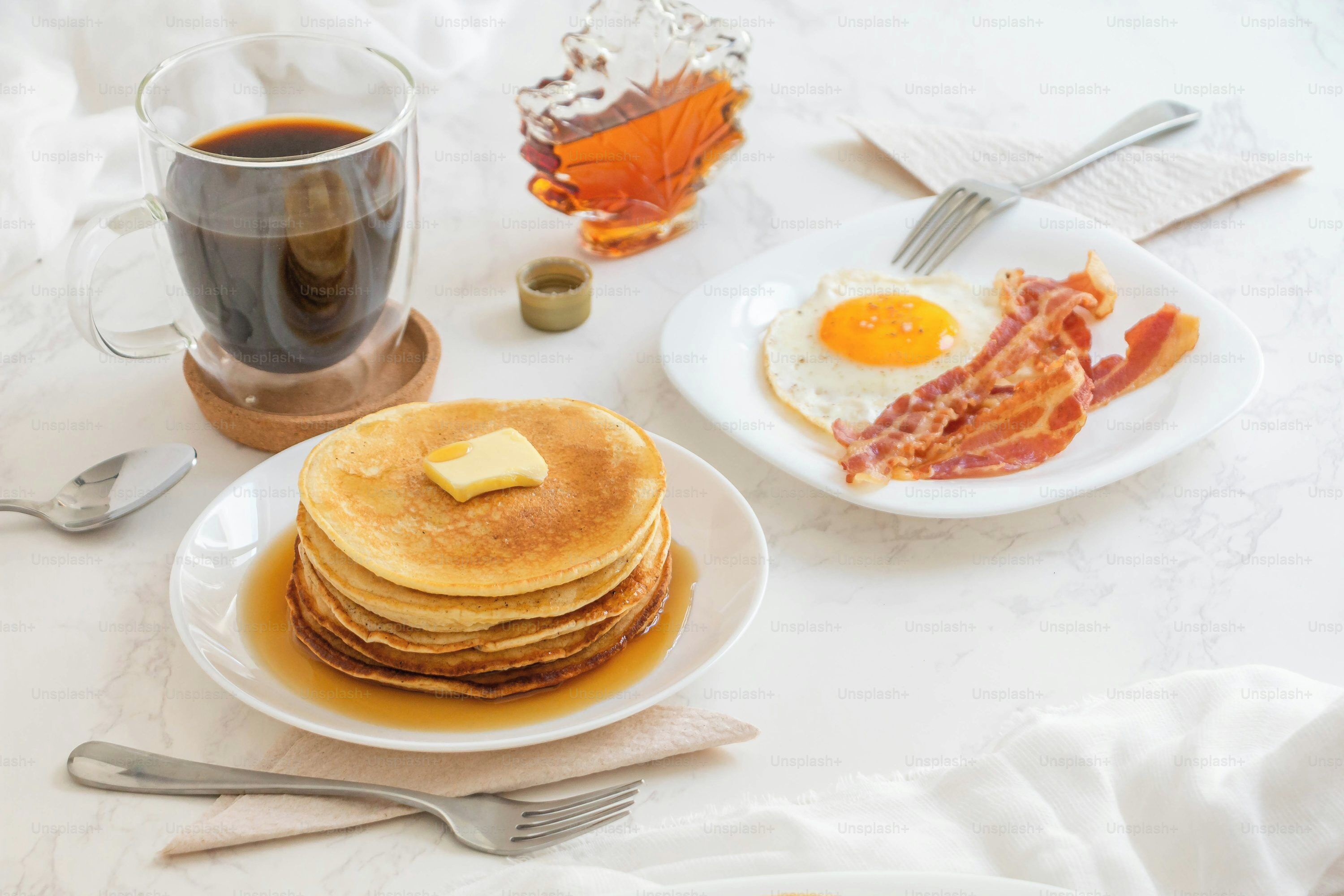 A breakfast setting with pancakes, a fried egg, bacon, a glass of coffee, and a bottle of syrup.