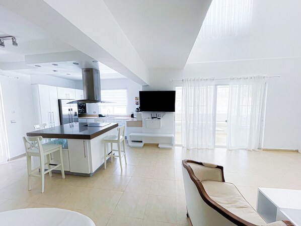 A modern kitchen with white cabinetry, a central island, and a built-in stove. There is a dining area with white chairs and a large window with sheer curtains. - BEACH FUN AND LIFE  (Punta Cana)