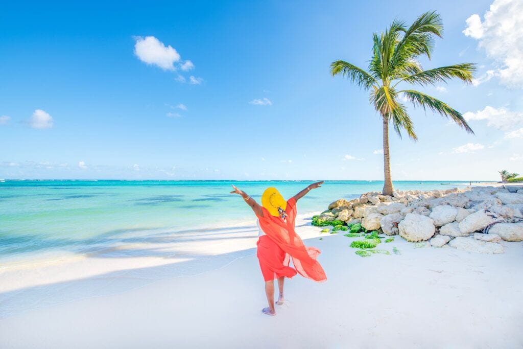 Eine Person in einem roten Kleid steht auf einem weißen Sandstrand, ein klarer, blauer Himmel und eine Palme sind zu sehen.