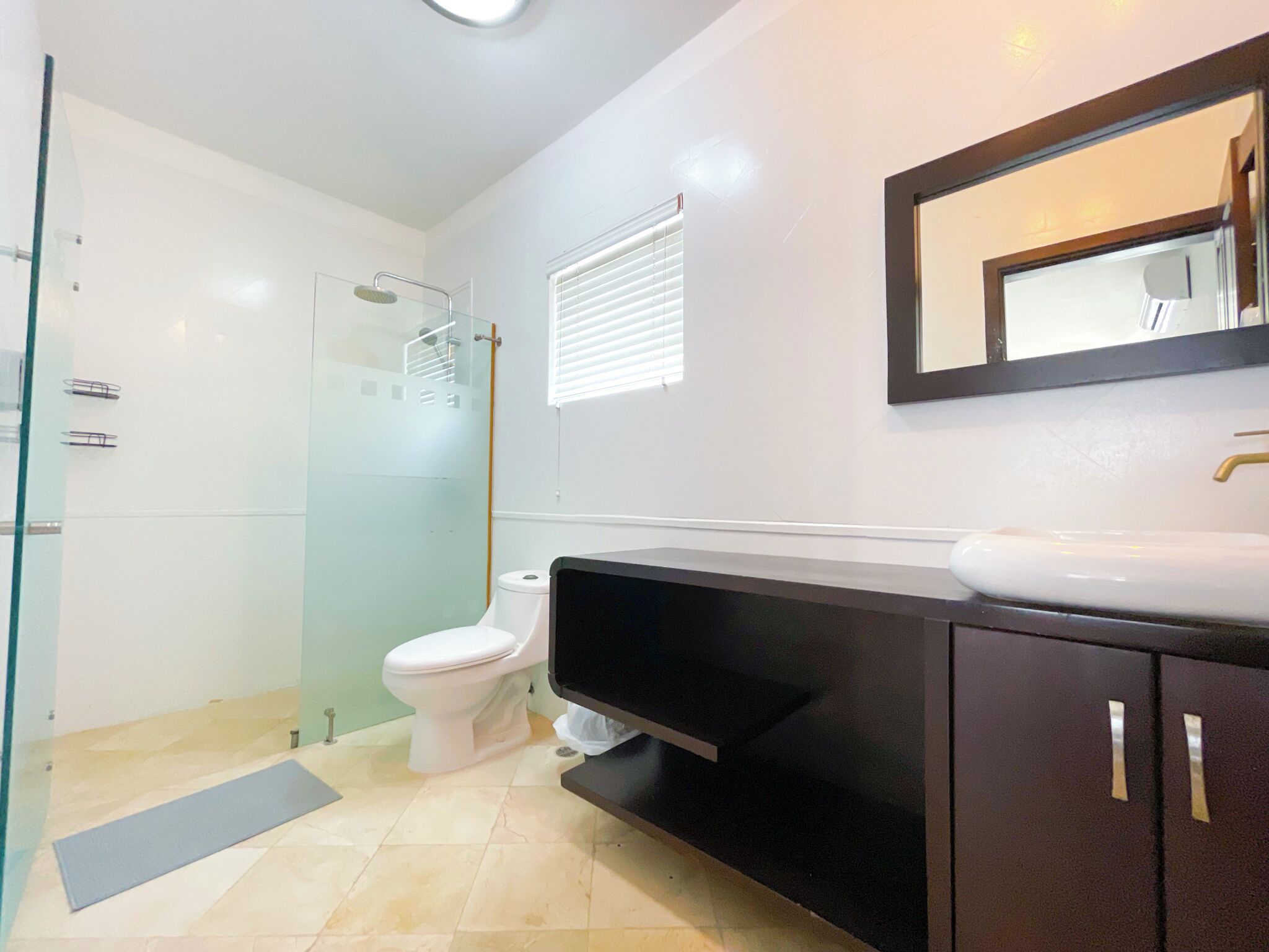A modern bathroom with a glass shower enclosure, a white toilet, a black vanity with a mirror, and a beige tiled floor.