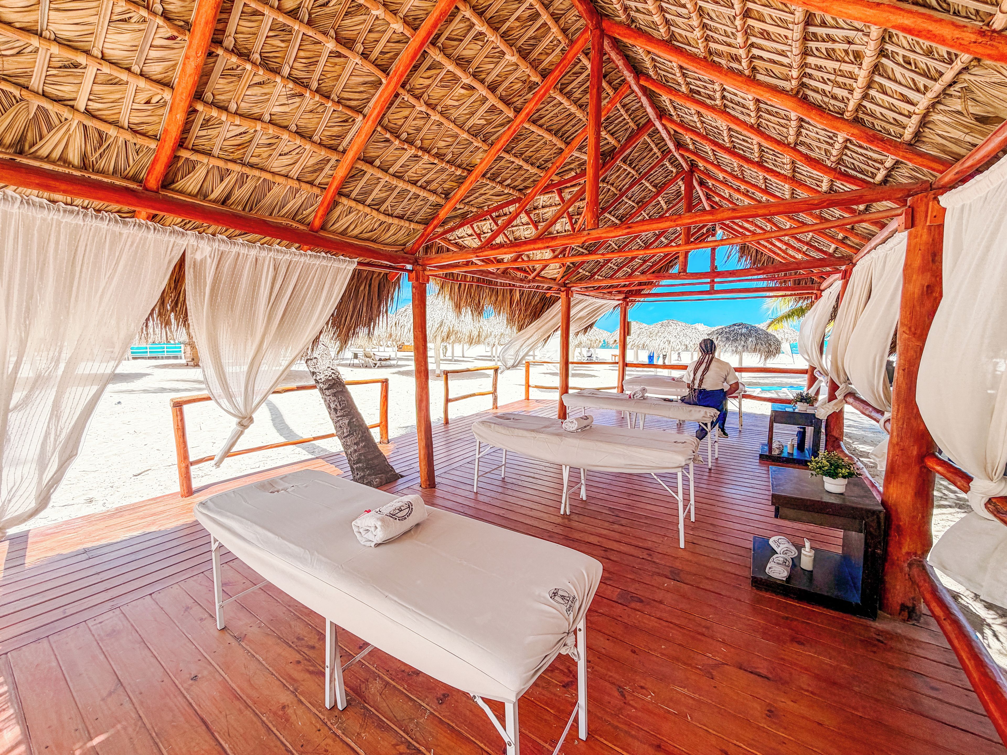 Un espacio de descanso frente a la playa, con piso de madera, sillones blancos y una estructura de techo de paja.