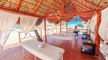 Un espacio de descanso frente a la playa, con piso de madera, sillones blancos y una estructura de techo de paja.