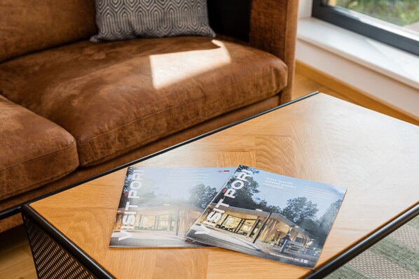 Una revista sobre una mesa de centro de cristal en un salón con un sofá marrón y una ventana al fondo.
