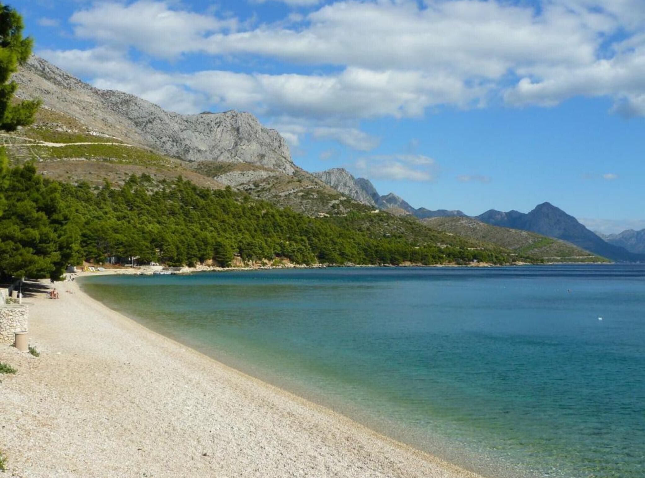 Una spiaggia con ciottoli, acqua cristallina e montagne sullo sfondo.